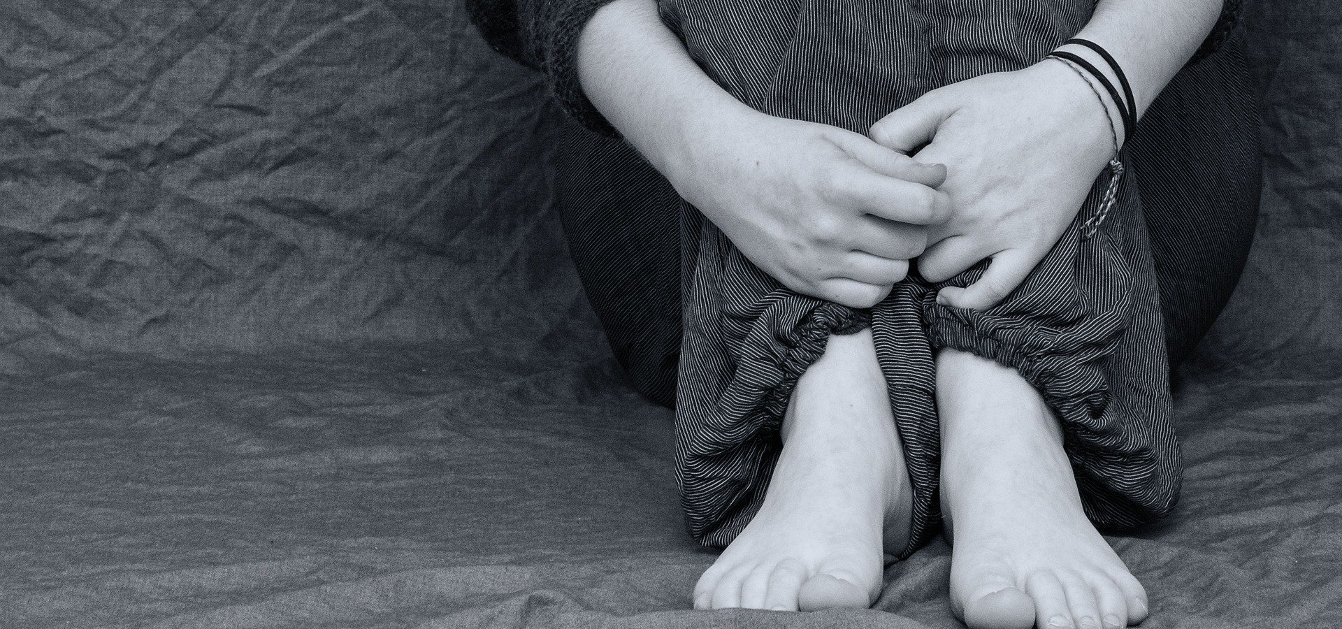 Zdjęcie przedstawia siedzącą kobietę. Widoczne są jej bose stopy i zgięte nogi, na których trzyma ręce.