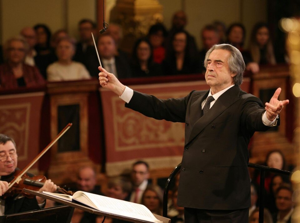 Na ilustracji widać dyrygenta, starszego mężczyznę, ubranego w garnitur, krawat i białą koszulę, który prowadzi orkiestrę podczas koncertu. W tle widać publiczność.