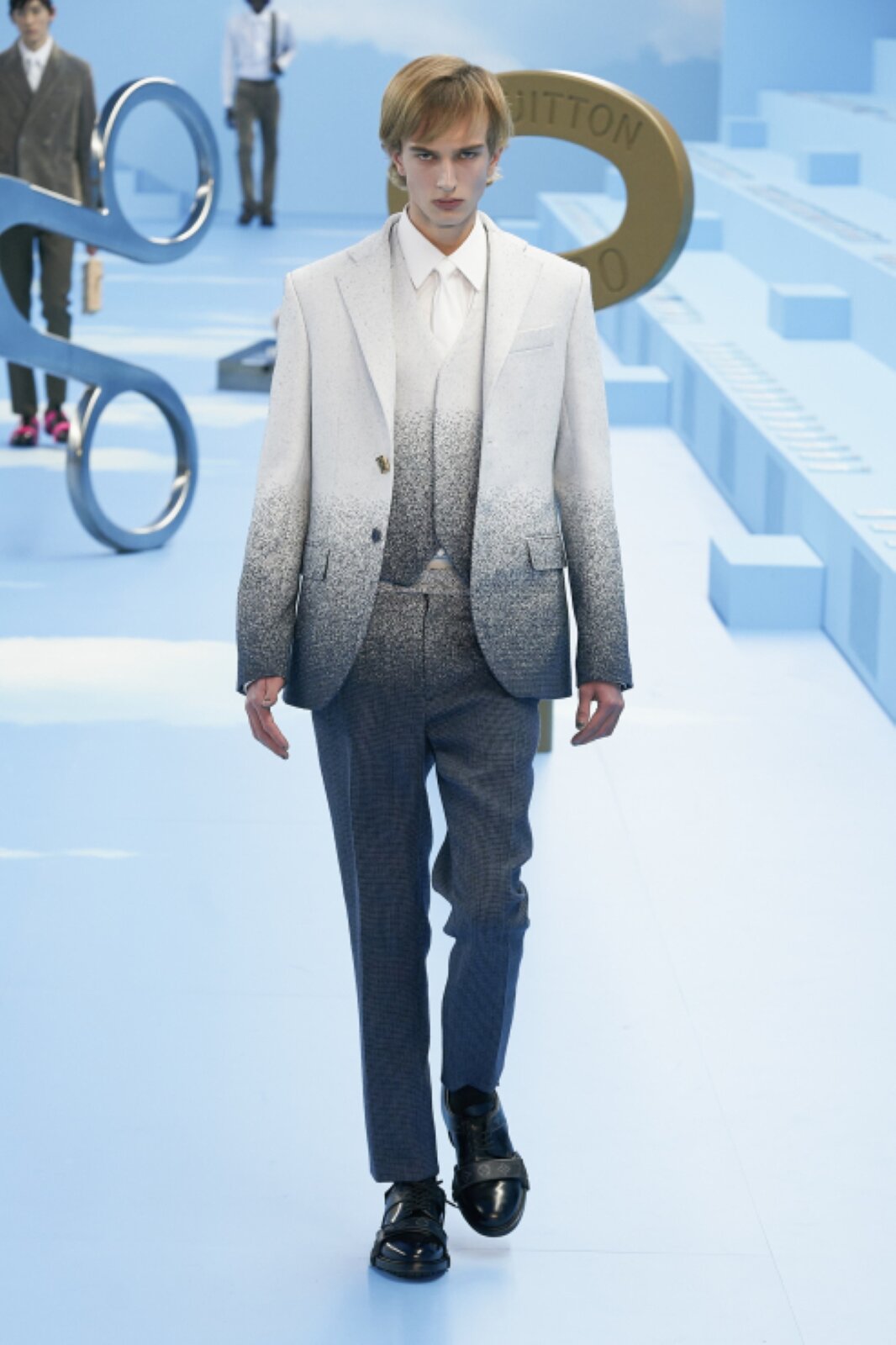 Fotografia przedstawia jasnowłosego młodego mężczyznę, który ma na sobie garnitur, który, od góry, w stylu „ombre” płynnie przechodzi z koloru białego do ciemnego granatu. Pod garniturem widoczna jest kamizelka w podobnym stylu i odcieniu oraz biała koszula i biały krawat. Kolor spodni to głównie ciemny granat, z elementami przejściowego zmieszania kolorów u góry.