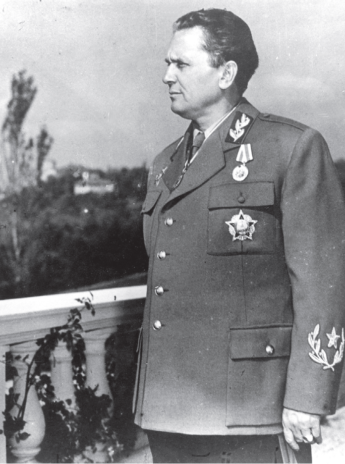 Na czarno‑białym zdjęciu znajduje się Josip Bros Tito. Ma on krótkie czarne włosy i jest ubrany w mundur do którego ma przypięte kilka orderów. Stoi on na tarasie i patrzy się w dal. w oddali widoczne są drzewa i nieliczne zabudowania.