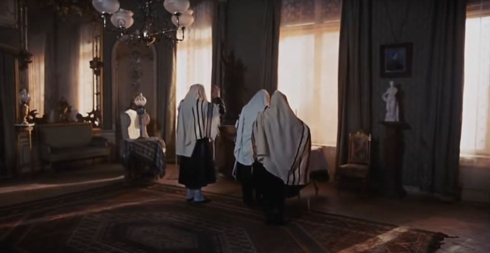 Zdjęcie przedstawia kadr filmowy, w którym ukazane są trzy postacie stojące tyłem do widza. Ubrane są w jasne chusty, a pierwsza z lewej unosi prawą rękę do góry. Postacie zwrócone są w stronę dużych okien z firanami i zielonymi kotarami. Pomiędzy oknami, przy ścianie, znajduje się biała figurka ustawiona na niewielkim postumencie.

Po lewej stronie na ścianie zawieszone jest duże lustro, pod którym stoi zielona sofa. Na podłodze leży ciemny, rozłożysty dywan. Na suficie widoczna jest lampa z czterema szklanymi abażurami. Obok postaci, po lewej stronie kadru, stoi fotel z wysokim oparciem, a przy nim niski stolik z lampą stołową.