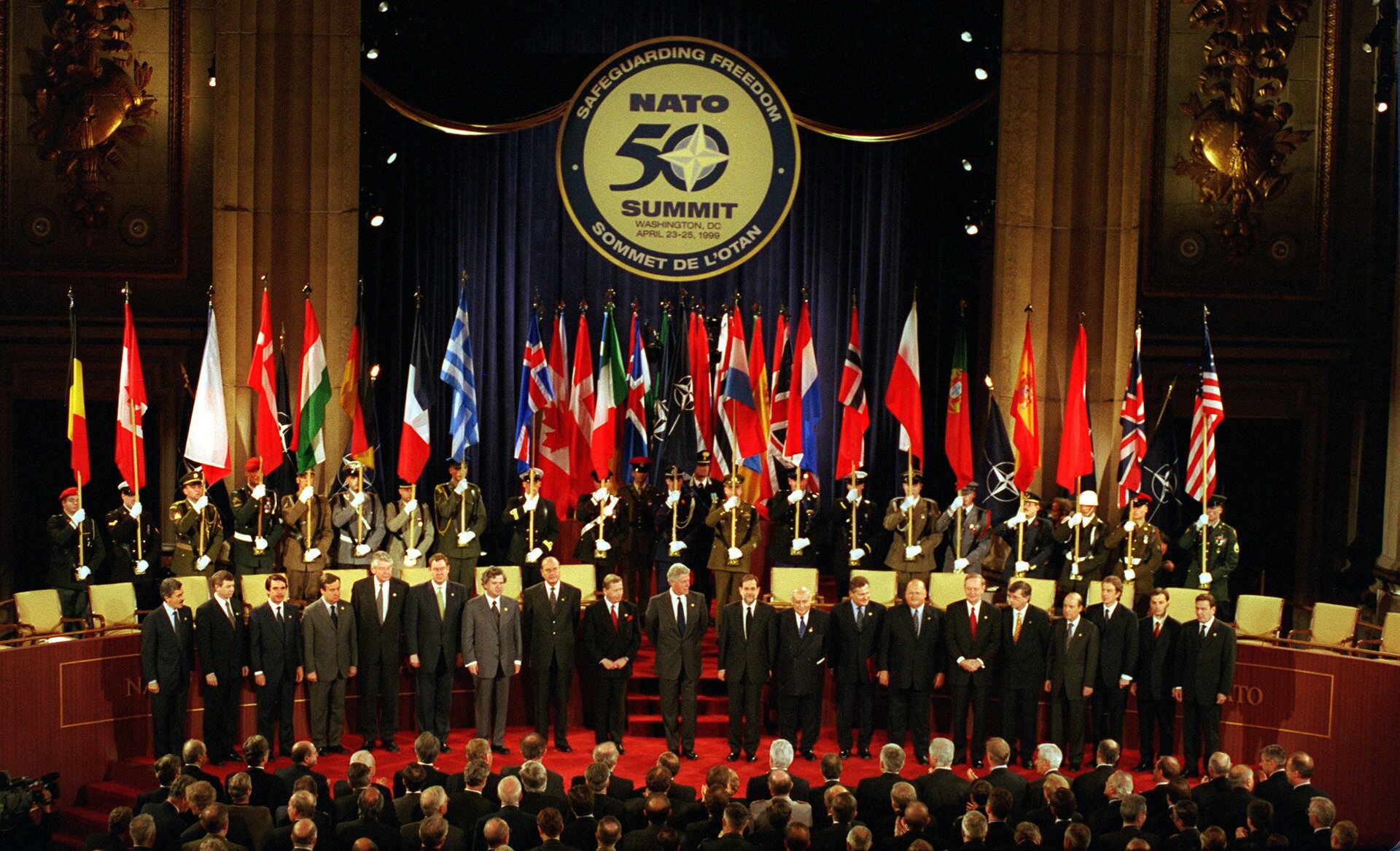 Fotografia barwna przedstawiająca Szczyt NATO w Waszyngtonie (1999). Z przodu ustawieni politycy a za nimi żołnierze trzymający flagi państw, z których pochodzą.