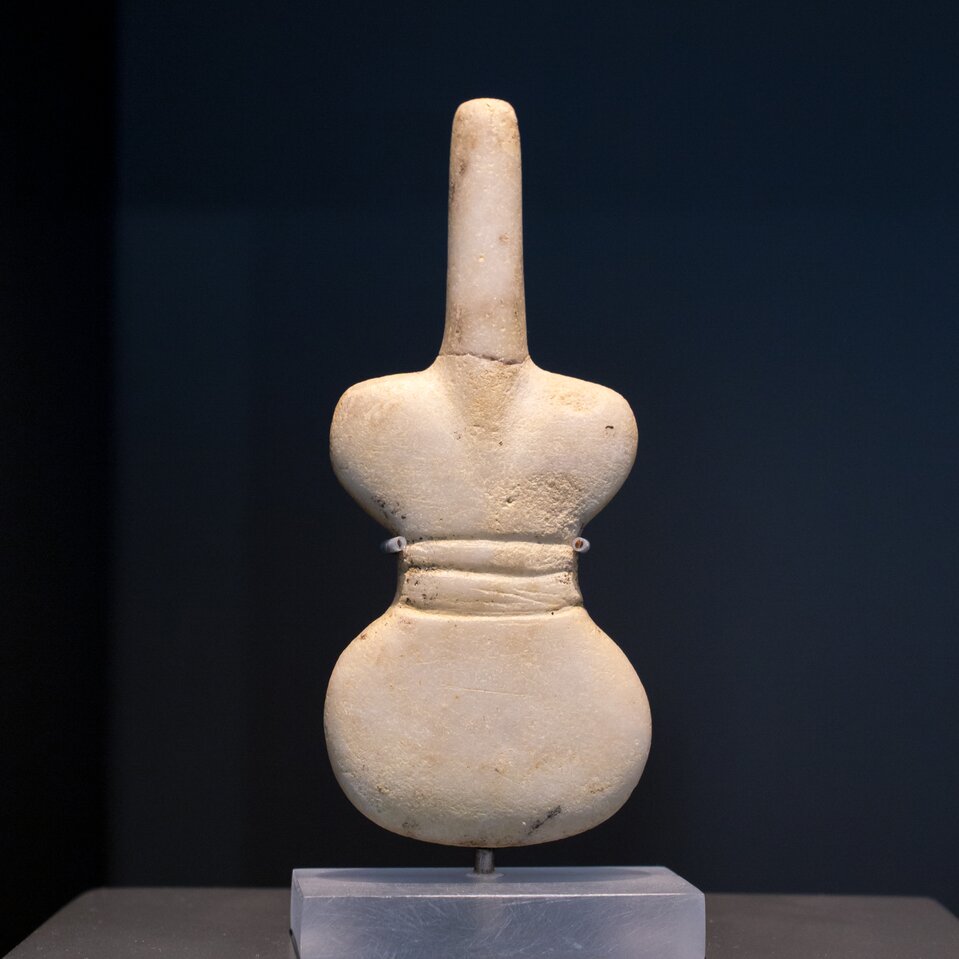 Ilustracja przedstawia figurkę idola. Ukazuje kobietę o uproszczonych kształtach. Postać przypomina skrzypce lub wiolonczelę. Figurka jest zredukowana   do podstawowych, geometrycznych zarysów.  Posiada rozszerzający się ku górze tors, wąską szyję, a zamiast typowej głowy ma wydłużony element pozbawiony detali i rysów twarzy. Figurka nie posiada kończyn. Figurka mieści się w dłoni i przypuszczalnie była używana w praktykach rytualnych lub grobowych.