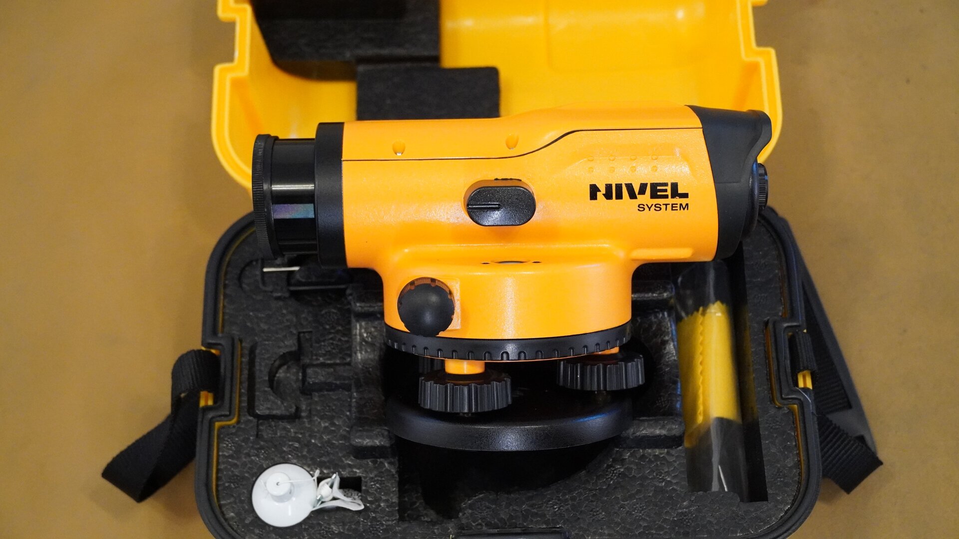 Na zdjęciu przedstawiono zbliżenie niwelatora na tle skrzynki, w której jest przechowywany. Instrument posiada czarną okrągłą podstawę – spodarkę, na niej znajdują się śruby, a na nich korpus z lunetą, która z jednej strony ma okular, a z drugiej obiektyw. Widoczne jest również jedno pokrętło. Korpus urządzenia jest w kolorze żółtym, a podstawa, śruby, pokrętła oraz elementy obudowy są czarne. 