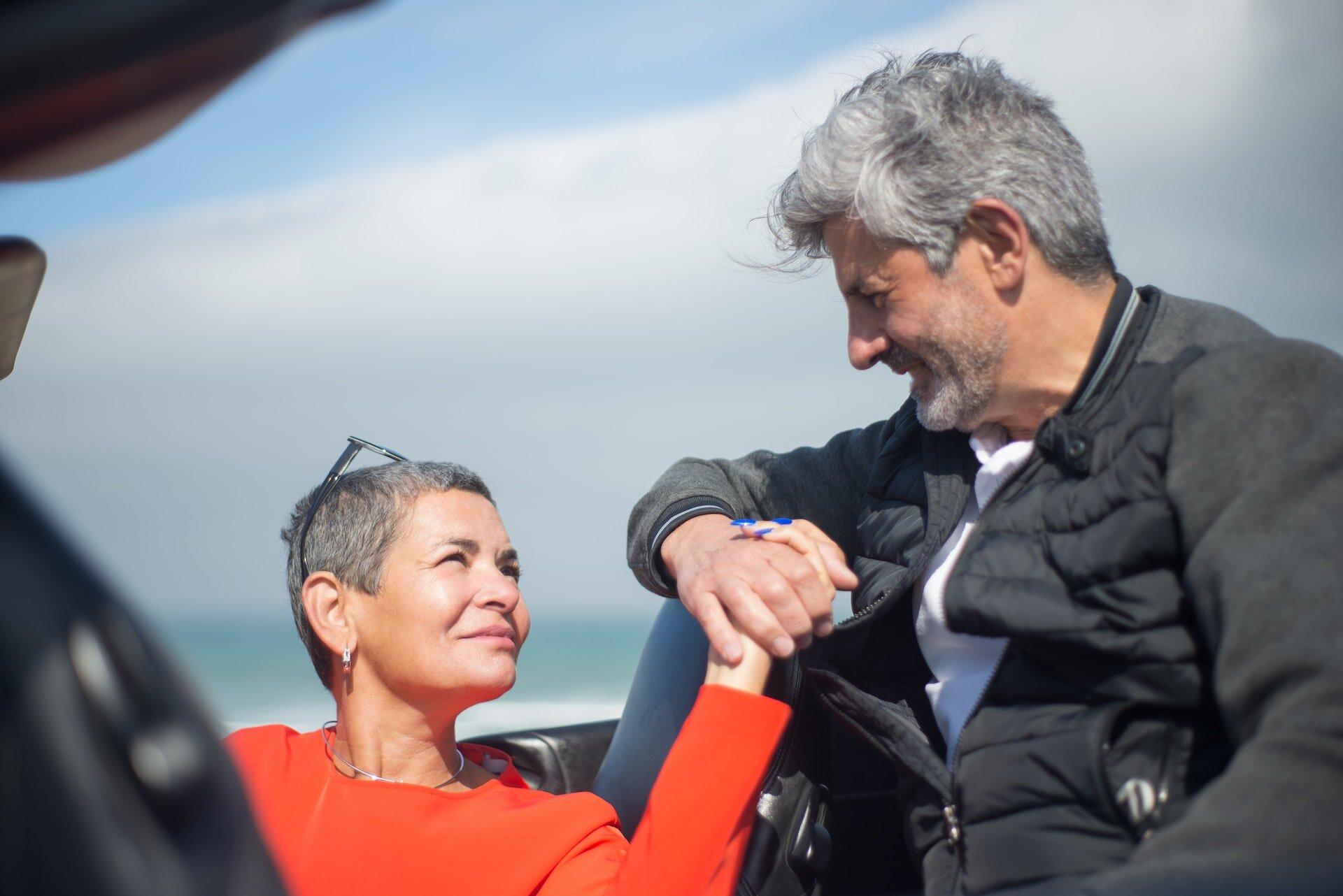 Zdjęcie przedstawia starszą parę trzymającą się za ręce. Kobieta ma na sobie pomarańczową bluzkę. Jej włosy są krótkie i siwe. Ma głowie ma okulary. Mężczyzna ma na sobie białą koszulkę i czarną bluzę. Ma siwe włosy i zarost.