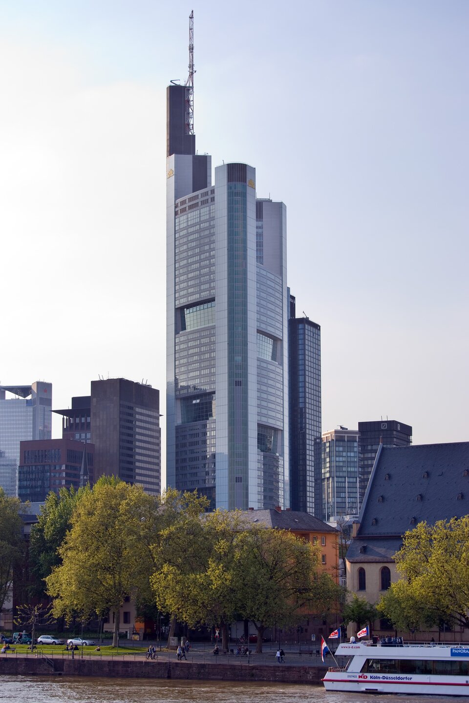 Ilustracja przedstawia Commerzbank Tower we Frankfurcie nad Menem w Niemczech. Jest to najwyższy wieżowiec w Europie. Budynek jest cały oszklony. Dookoła widzimy inne, niższe budynki oraz drzewa.
