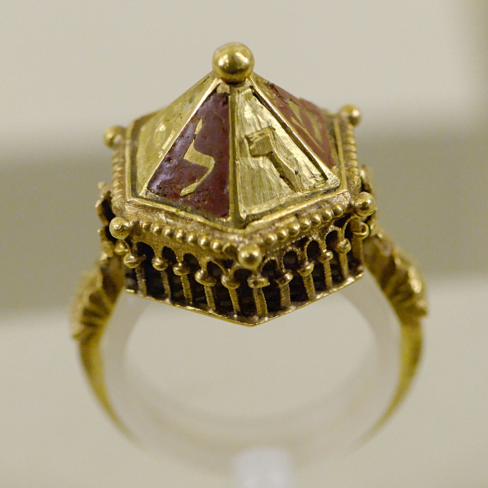 Ilustracja przedstawia złoty pierścień. Ma on stożkowaty kształt, jest bogato zdobiony po bokach, a na czubku zakończony małą kuleczką.