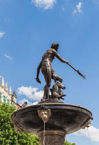 Fotografia przedstawia fontannę Neptuna. Figura boga wykonana jest z brązu, zaś czasza fontanny z czarnego marmuru. Bóg ukazany jest nago, w pozycji stojącej. W prawej ręce trzyma trójząb, w lewej muszlę. U jego stóp znajdują się dwa hipokampy. Jeden z nich owija ogon wokół uniesionej nogi boga. Do czaszy fontanny przymocowane są marmurowe lwie głowy, z których wypływa woda.