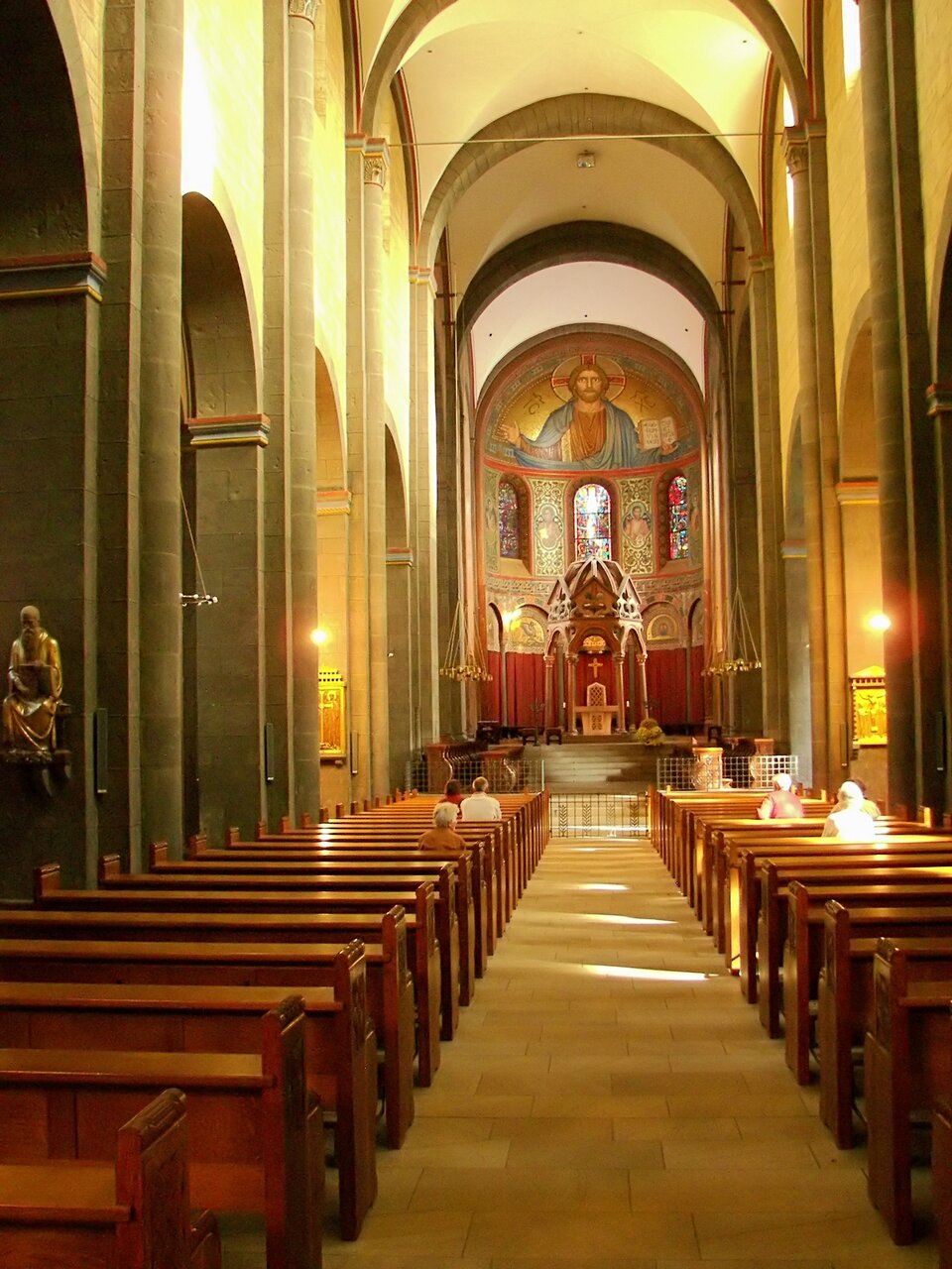 Ilustracja o kształcie pionowego prostokąta przedstawia wnętrze kościoła Maria Laach. Fotografia ukazuje nawę główną z widokiem na prezbiterium i apsydę. Jest sklepiona krzyżowo. Po bokach znajdują się arkadowe ściany oddzielająca nawę główną od bocznych z półkolistymi łukami wspartymi na filarach. Wzdłuż nawy ustawione są drewniane ławki, a pośrodku wolna przestrzeń. Na zakończeniu nawy, w sklepieniu nad prezbiterium, znajduje się malowidło z Chrystusem Pantokratorem. 