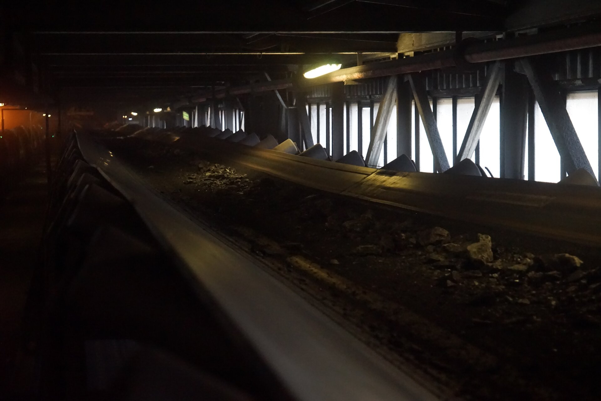 Na zdjęciu widoczny jest przenośnik taśmowy transportujący węgiel. Na czarnej taśmie widoczne są drobiny węgla. Taśma osadzona jest na rolkach o kształcie walca. Taśmociąg znajduje się w zamkniętym budynku. Na prawej ścianie widoczne są okna.