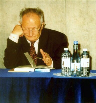 Zdjęcie przedstawia mężczyznę, który siedzi przy stole przykrytym niebieskim obrusem. Mężczyzna jest siwy i ma założone okulary. Jest ubrany w czarny garnitur, białą koszulę i bordowy krawat. Mężczyzna opiera głowę o prawą dłoń. Lewą ręką przekartkowuje książkę, która leży przed nim na stole. Z prawej strony zdjęcia stoją trzy butelki wody – dwie o podstawie kwadratu, jedna koła. Ściana za mężczyzną jest w kolorze kremowym.