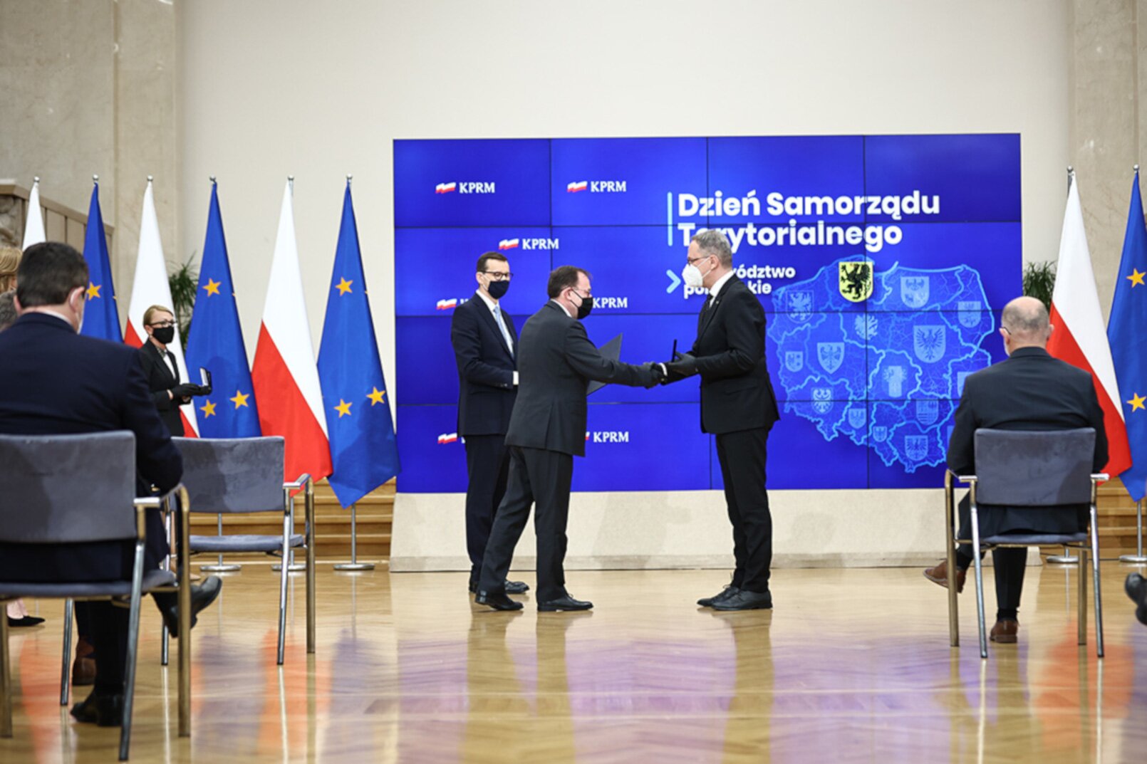 Zdjęcie przedstawia trzech mężczyzn w garniturach stojących na środku sali. Dwóch jest naprzeciwko siebie i podaje sobie ręce. Trzeci stoi za nimi