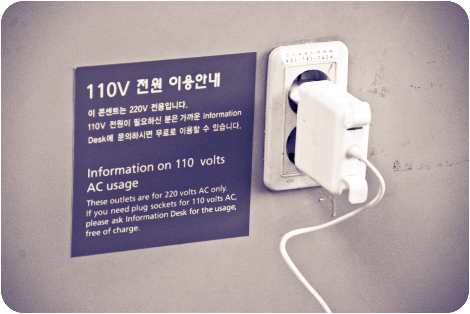 Ilustracja przedstawia wtyczkę białą wetkniętą w podwójne białe gniazdo elektryczne. Od wtyczki odchodzi biały kabel. Obok tablica w kolorze fioletowym z napisami w języku japońskim i angielskim. Tło szare.