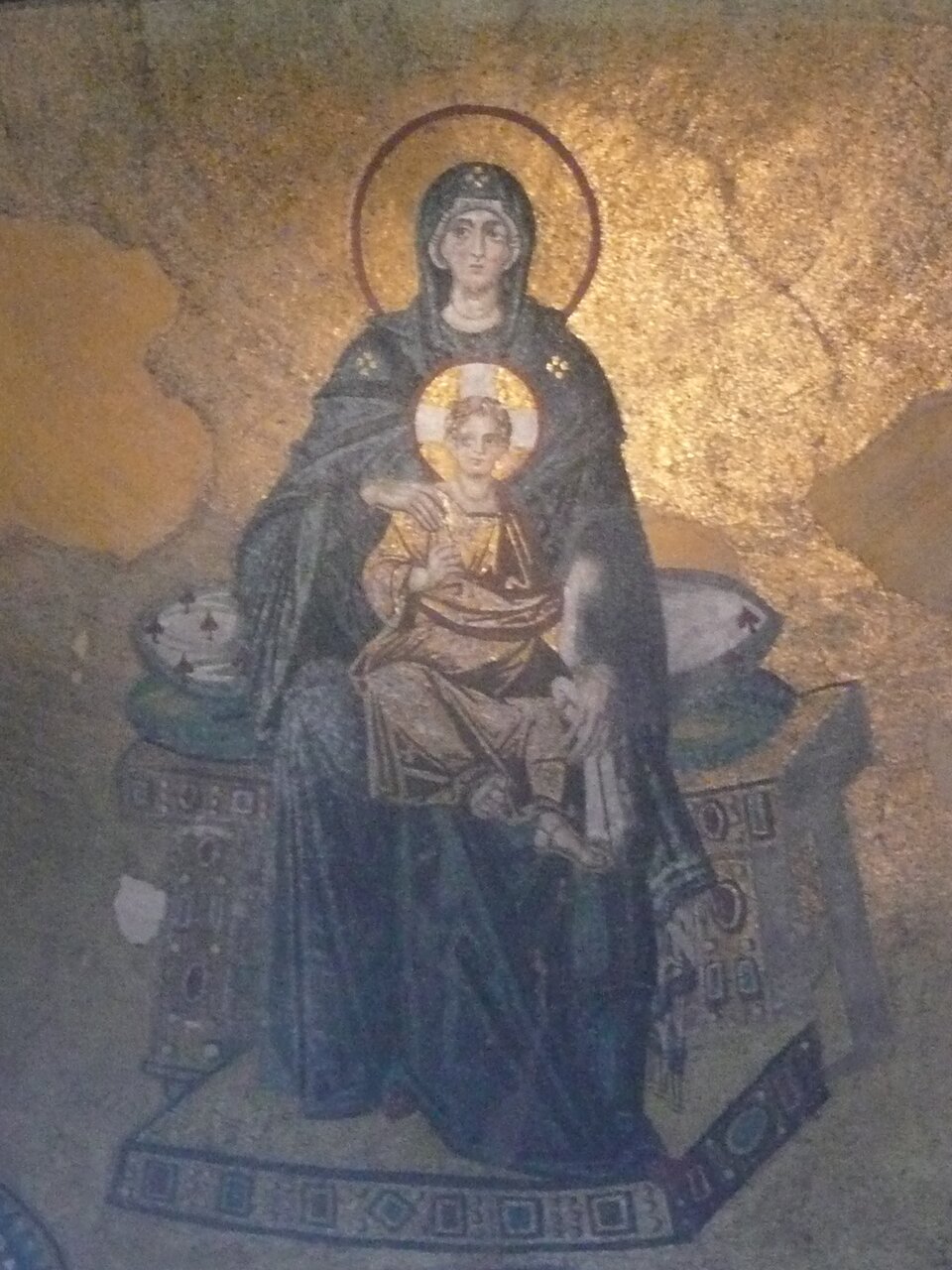 Mozaika bizantyjska przedstawiająca Matkę Bożą tronującą z Dzieciątkiem na kolanach: obie postacie mają aureole i ukazane są frontalnie na złotym tle. Maryja ubrana jest w ciemny płaszcz, podtrzymuje Jezusa ubranego w jasną szatę, a całość ma charakter hieratyczny i symetryczny, z widocznymi śladami starzenia i ubytkami mozaiki.