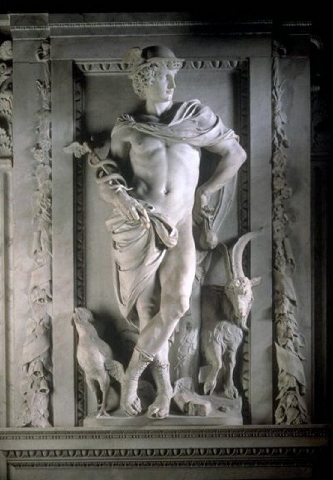 Ilustracja przedstawia rzeźbę Merkurego. Bóg ukazany jest z charakterystycznym kaduceuszem w dłoni, na głowie ma podróżny petasos, a na stopach sandały ze skrzydłami. Spod kapelusza wystają kręcone włosy. Cała sylwetka ustawiona jest esowato, nogi poniżej kolan są skrzyżowane. Płaszcz spowija ramiona boga oraz osłania jego podbrzusze. U stóp Merkurego stoi kozioł z dużymi rogami oraz kogut.