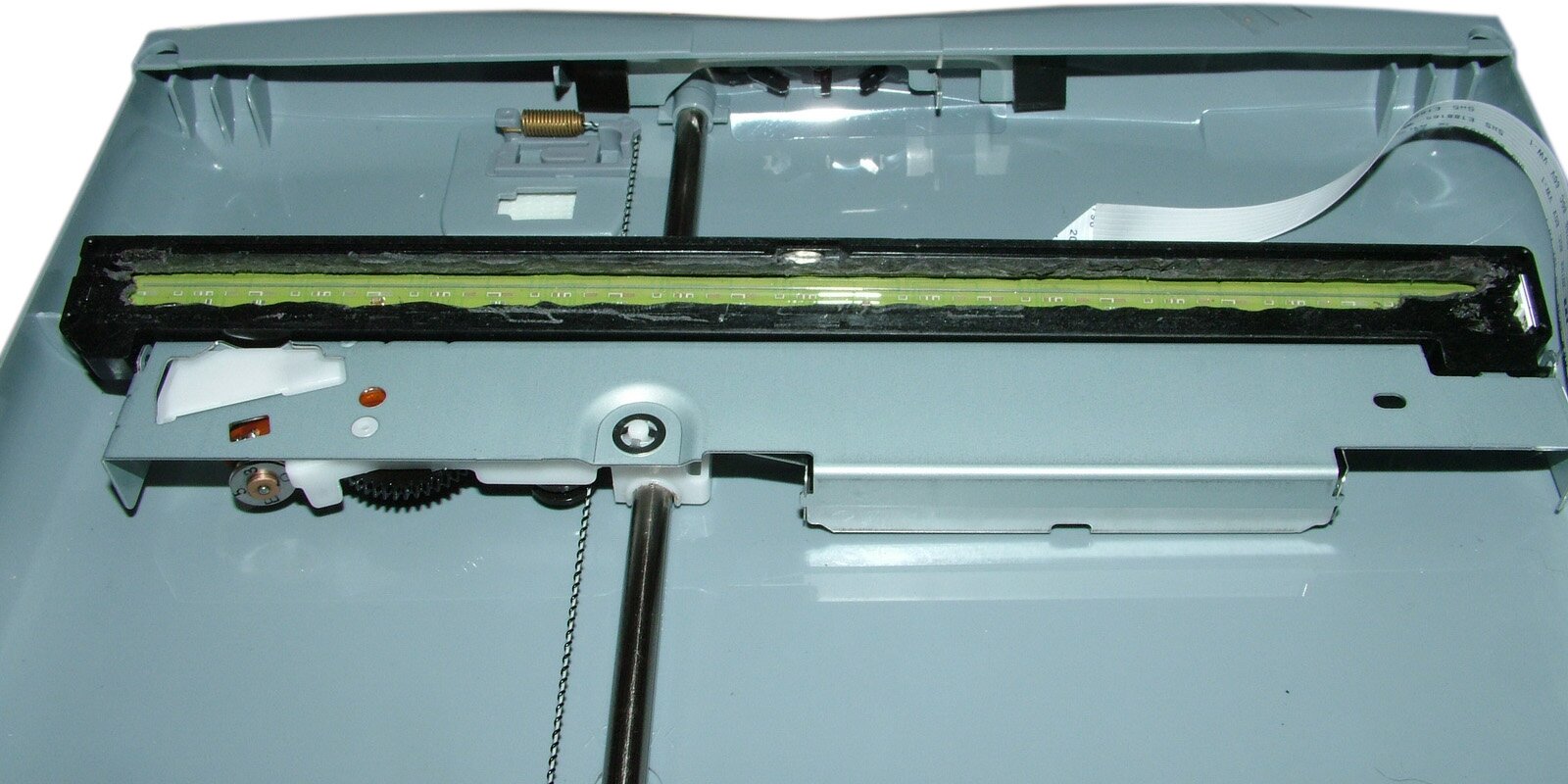 Zdjęcie przedstawia czujnik światłoczuły w skanerze płaskim. Widoczna jest część wnętrza skaner z obudową, różnymi śrubkami i kółkami oraz  poziomym czarnym elementem, biegnącym wzdłuż, w którym osadzona jest zielona taśma.