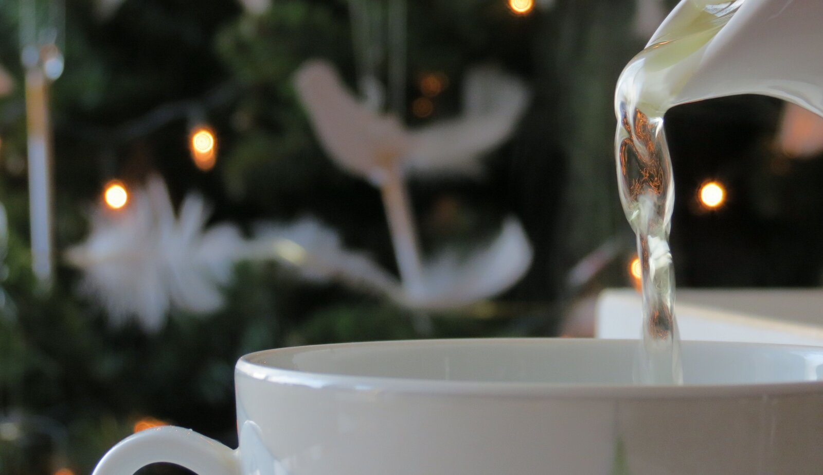 Kliknij żeby powiększyć Fotografia przedstawia rant filiżanki, do której jest wlewana woda. Woda nalewana jest z dzbanka, którego dzióbek widać w prawym górnym rogu zdjęcia. W tle widać niewyraźną choinkę bożonarodzeniową.