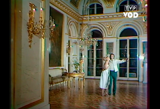 Fotografia przedstawia wnętrze pałacowej komnaty. Pośrodku stoi para młodych ludzi. Mężczyzna ma krótkie, ciemne włosy i bokobrody. Postać ukazuje w całości owalną twarz. Jest ubrany w koszulę, marynarkę i spodnie. Mężczyzna wysuwa do przodu lewą rękę. Prawą ręką obejmuje kobietę. Postać ukazuje prawy profil owalnej twarzy. Jej czoło jest przykryte grzywką. Jej długie włosy są upięte z tyłu głowy. Fryzura jest ozdobiona wąskimi wstążkami. Opiera głowę o ramię mężczyzny. Jest ubrana w obcisłą, długą sukienkę bez rękawów. Za ich plecami ukazane jest wysokie pomieszczenie bogato zdobione złotymi detalami. Przy ścianie po lewej stronie stoi kanapa i stolik. Nad kanapą wisi duży obraz oprawiony w szeroką ramę. U sufitu wisi kryształowy żyrandol. Okna są wysokie.