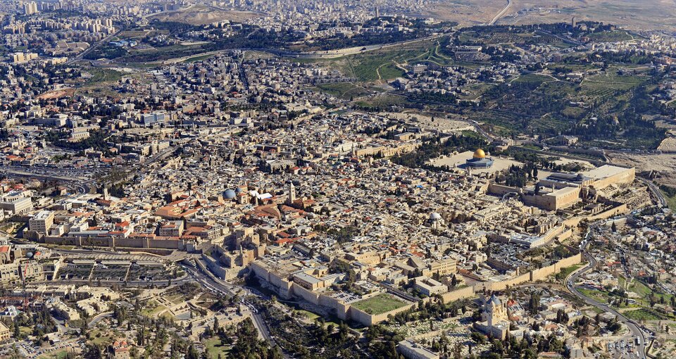 Zdjęcie przedstawia panoramę Starego Miasta w Jerozolimie z lotu ptaka. W dole widoczne liczne budynki z czerwonymi dachami. Na dalszym planie pusty plac z charakterystycznym błękitnym meczetem ze złotym dachem – Kopułą na Skale. 