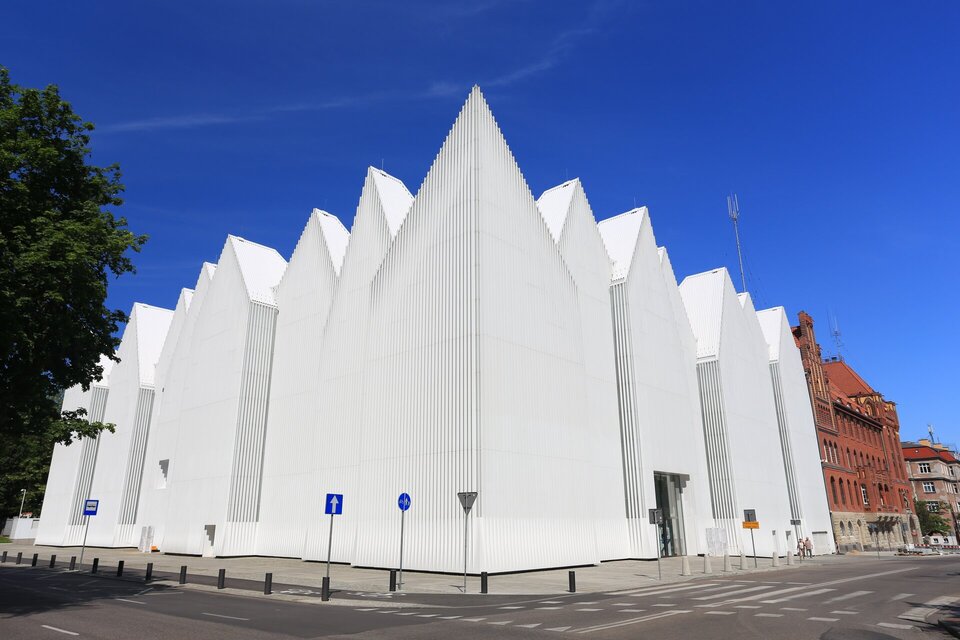 Ilustracja  przedstawia filharmonię im. Mieczysława Karłowicza w Szczecinie, zaprojektowaną przez barcelońskich architektów Estudio Barozzi Veiga. Znajduje się na zbiegu ulic. Bryła obiektu jest strzelista, nawiązuje strukturą do sąsiednich szczytów kamienic. Wyróżnia się białą fasadą, której elementy naprzemiennie wysuwają się lub cofają. Uhonorowana wieloma nagrodami architektonicznymi filharmonia szczecińska inspirowana jest bryłą góry lodowej. Koszt jej budowy to blisko 100 milionów złotych. Budowla jest nowoczesną ikoną Polski w świecie architektury.
