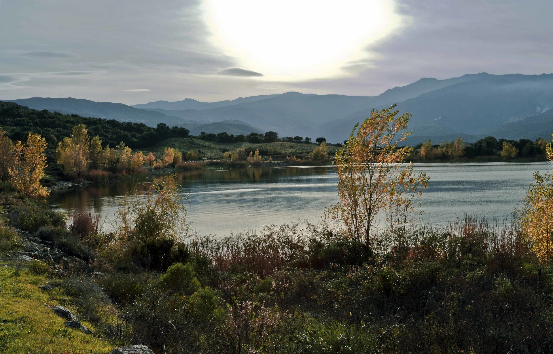 Fotografia prezentuje jezioro, wokół którego rosną różne rośliny oraz drzewa. W tle widoczne łańcuchy górskie. Krajobraz jesienny. Uchwycono zachodzące słońce. Niebo szare.