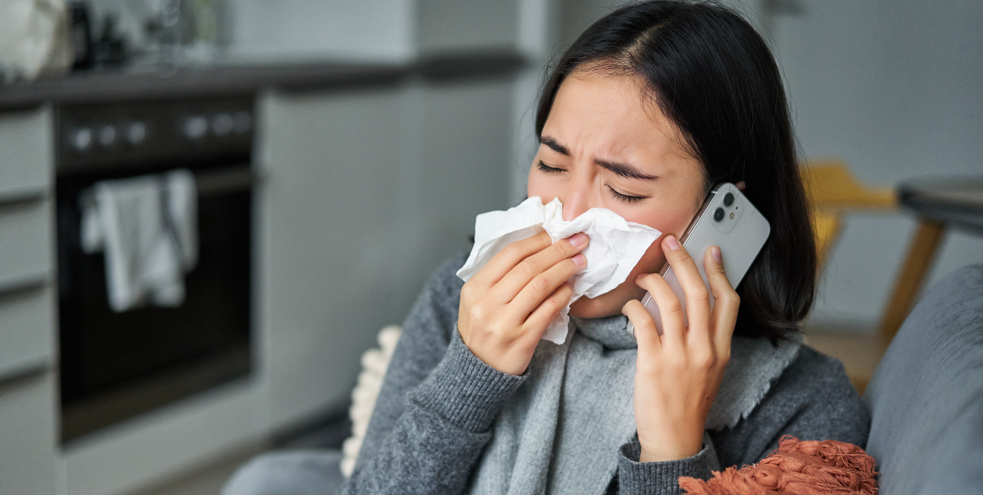 Ilustracja przedstawia kobietę z wypisanym na twarzy grymasem cierpienia. Kobieta wyciera nos chusteczką higieniczną. Do ucha przyłożony ma telefon.