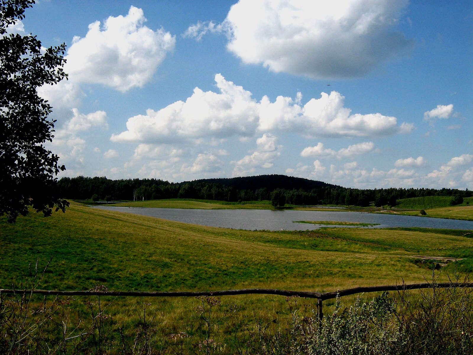 Zdjęcie ukazuje trawiasty teren, na którym znajduję się zbiornik wodny. Na pierwszym planie widać drewnianą barierkę. W tle ukazane są zalesione wzgórza. 