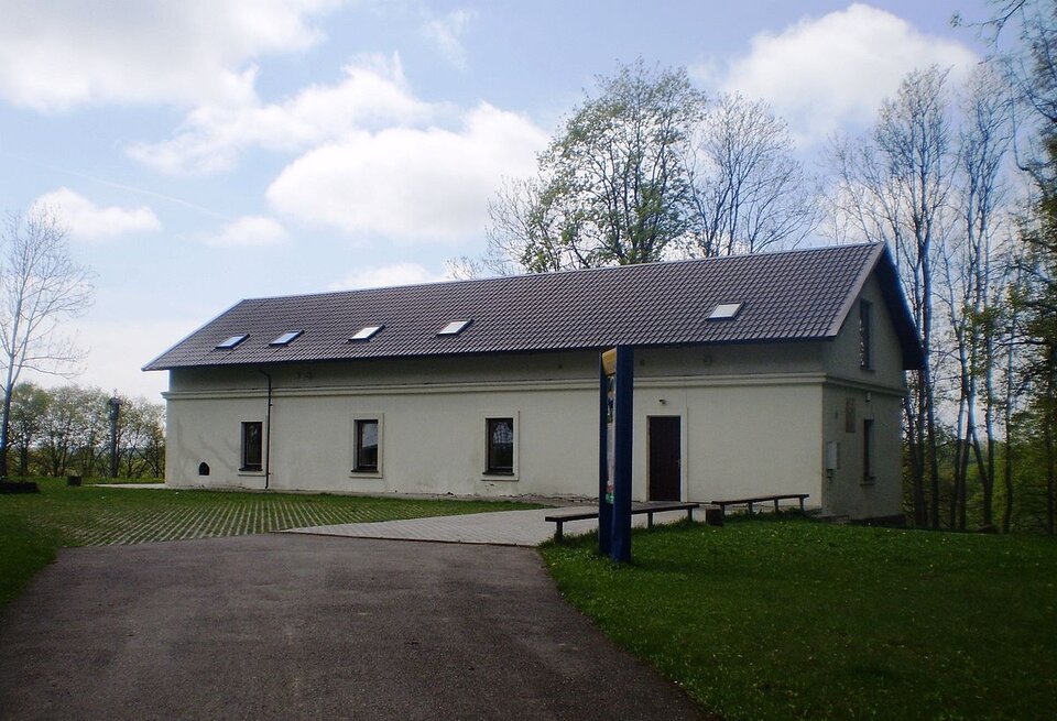 Zdjęcie przedstawia stojący między drzewami budynek gospodarczy. Budynek jest parterowy, w białym kolorze ze spadzistym dachem.