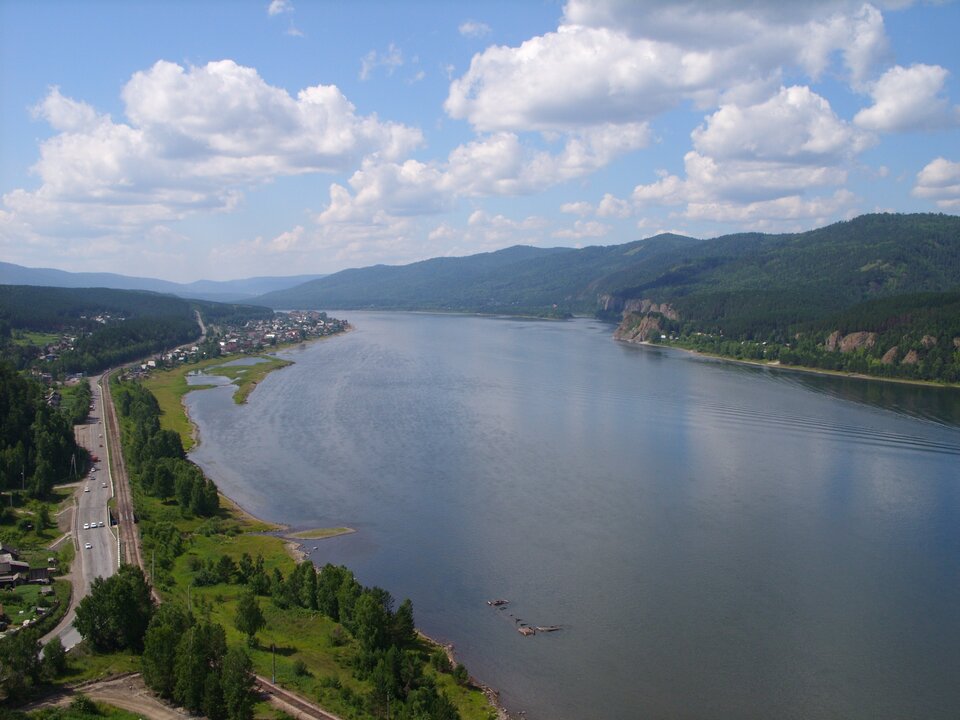 Zdjęcie przedstawia rzekę. Po lewej stronie widać zabudowania i drogę, po prawej – górski brzeg. Na horyzoncie wzgórza pokryte lasem i błękitne niebo z chmurami. Rzeka płynie szerokim, łagodnym korytem, otoczona jest zalesionymi wzgórzami.