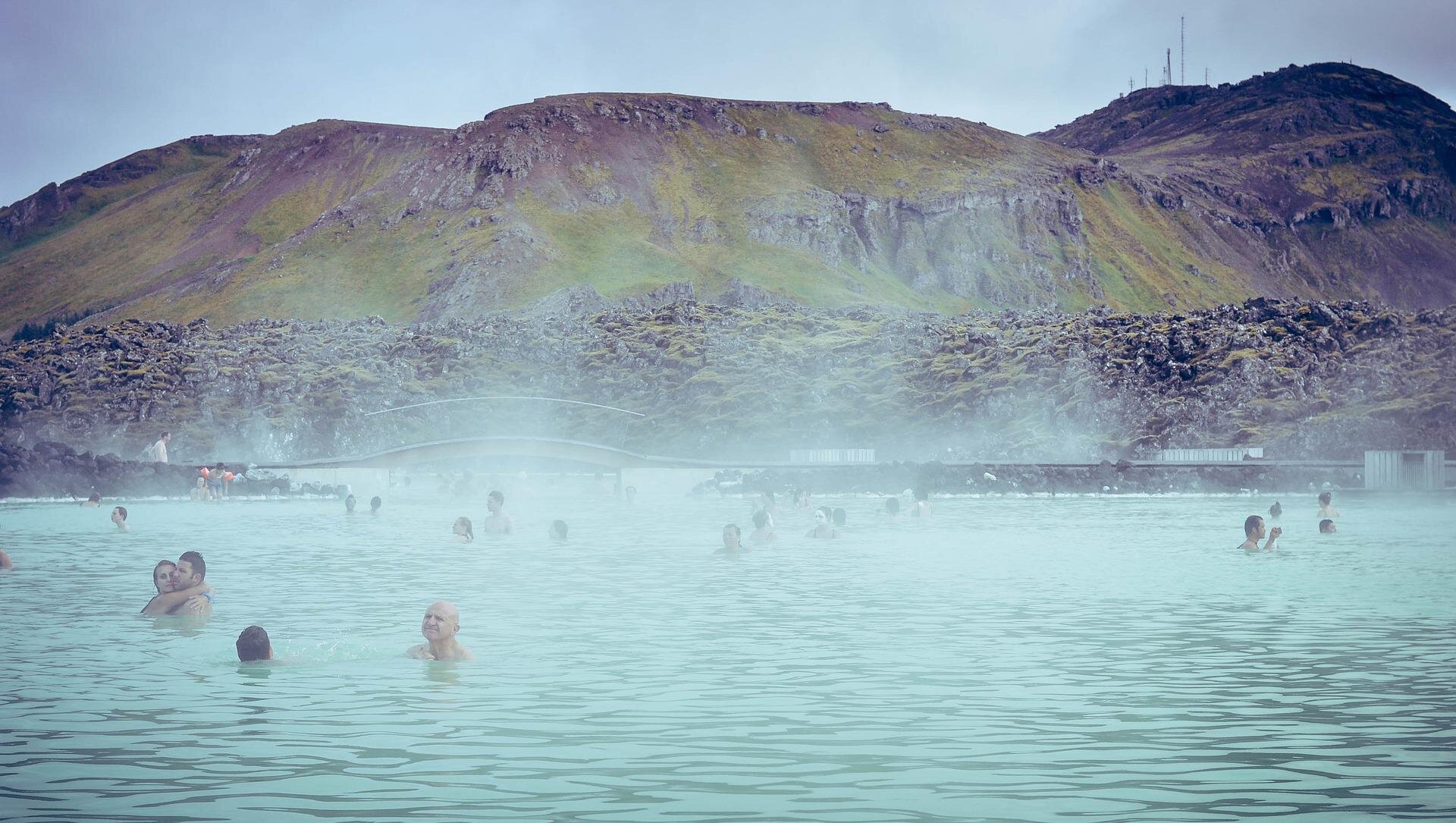 Zdjęcie przedstawia Błękitną Lagunę na Islandii. Nad powierzchnią wody unosi się gęsta para, na pierwszym planie widoczni są kąpiący się w wodzie ludzie. W tle za nimi znajduje się górzyste wybrzeże. Niebo na zdjęciu jest pochmurne, w szaroniebieskim kolorze.