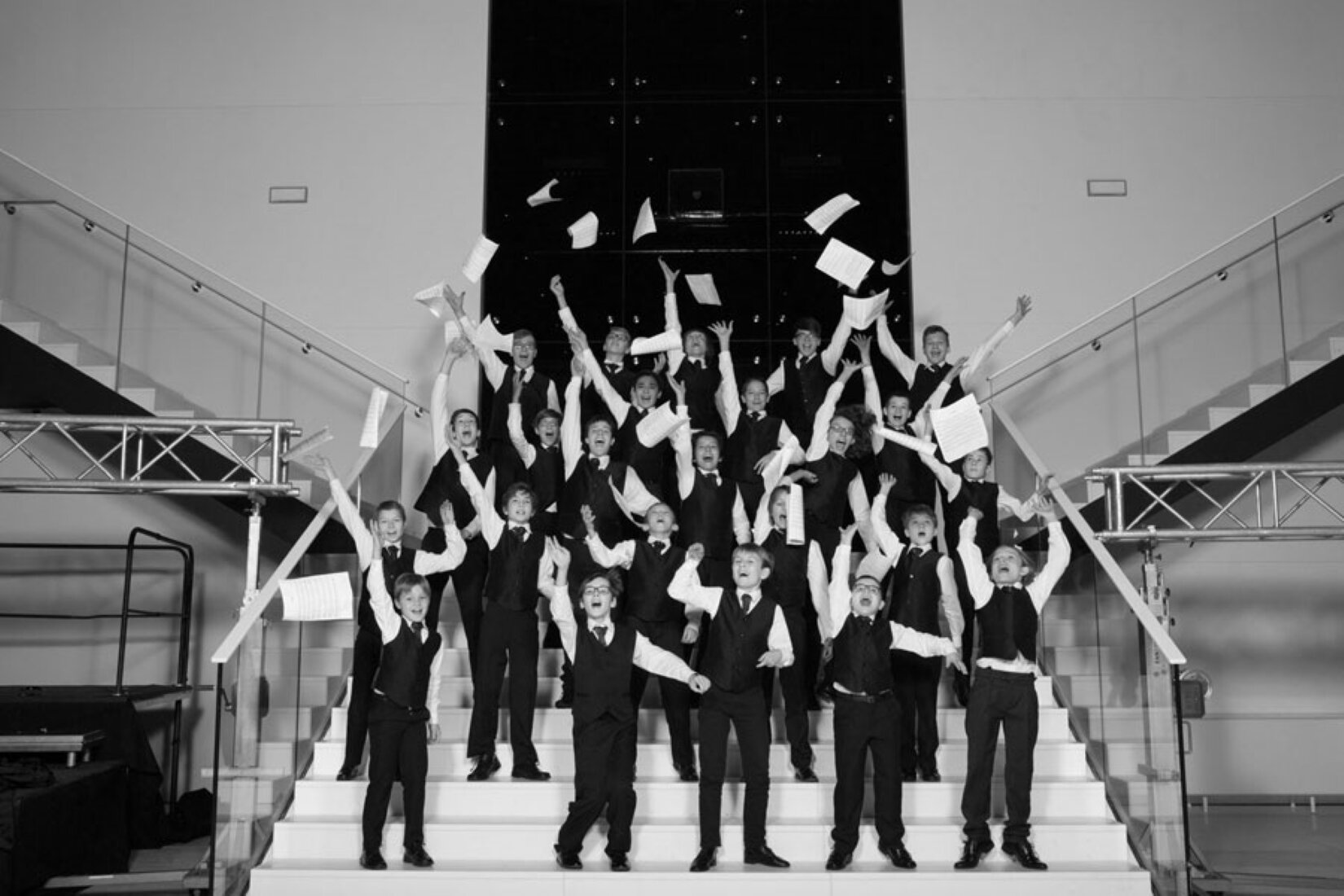 Czarno‑biała fotografia przedstawia chór chłopięcy Narodowego Forum Muzyki imienia Witolda Lutosławskiego we Wrocławiu. Chłopcy stoją na schodach i rzucają kartkami. Ubrani są w czarne spodnie garniturowe, białe koszule i czarne kamizelki, a na ich szyjach widoczne są krawaty. 
