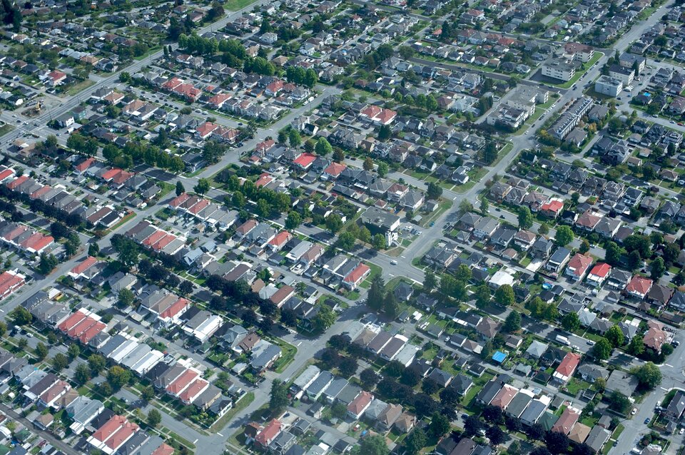 Zdjęcie przedstawia suburbia z lotu ptaka. Są to niewielkiej wielkości domy mieszkalne umiejscowione ściśle obok siebie z małymi podwórkami. Są one położone pomiędzy regularną siatką wyasfaltowanych ulic.