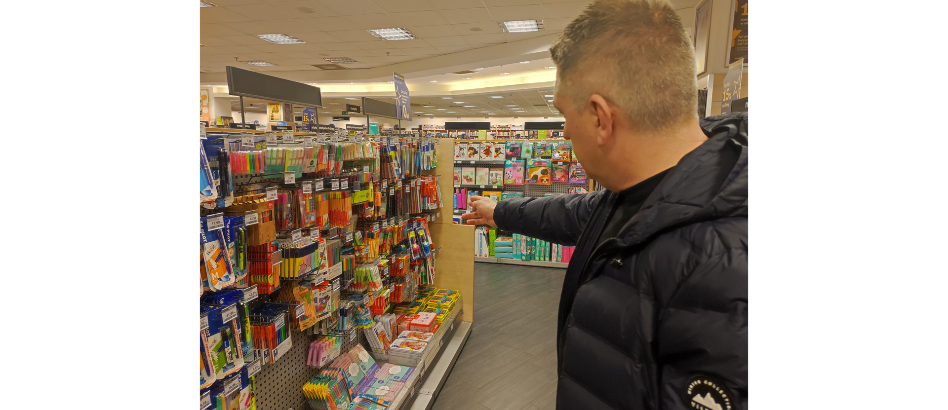 Zdjęcie przedstawia mężczyznę wskazującego ręką na regał z artykułami papierniczymi.