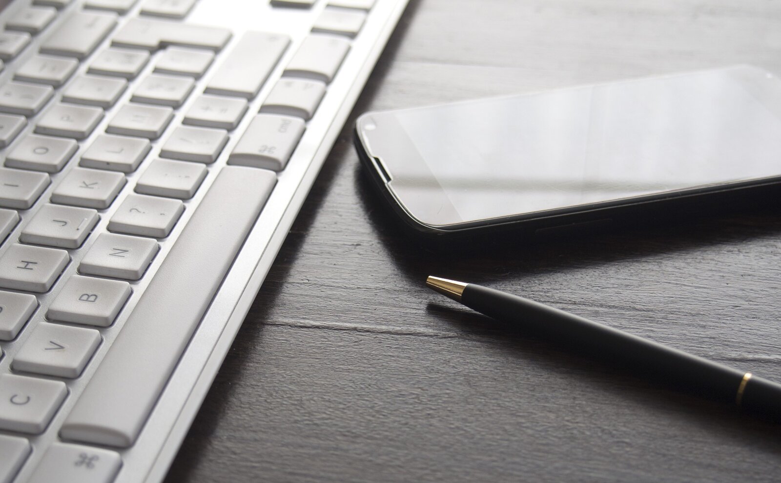 Fotografia przedstawia fragment białej klawiatury komputerowej. Na prawo od niej leży telefon dotykowy oraz wyłączony długopis.