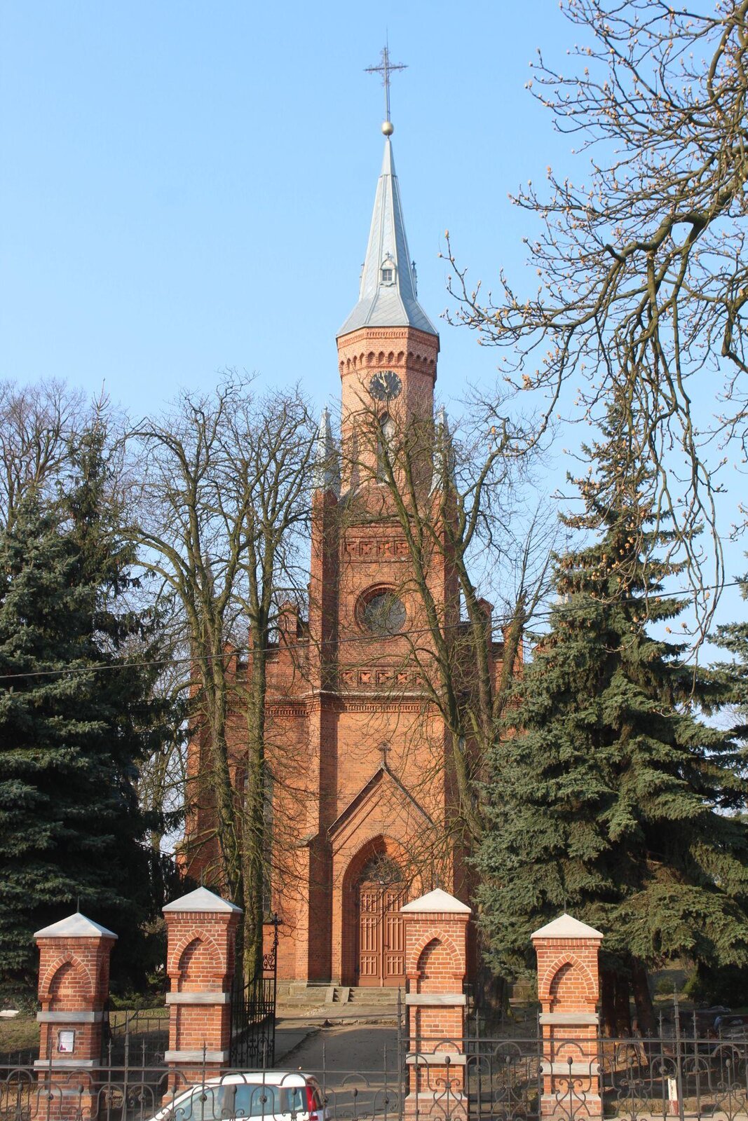 Zdjęcie przedstawia ceglany, wysoki kościół, którego wejście zakończone jest wysoką wieżą z zegarem i krzyżem na czubku. Przed kościołem rosną wysokie choinki i bezlistne drzewa. Teren kościoła jest ogrodzony płotem, którego część wykonana jest z czerwonych cegieł. Niebo jest błękitne, bezchmurne. Zdjęcie zostało wykonane w świetle dziennym.