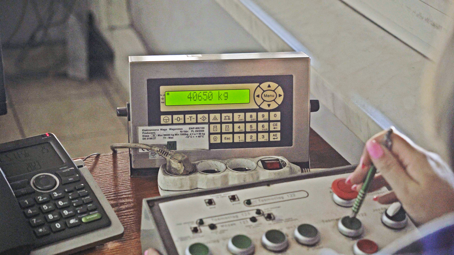 Na zdjęciu widoczne jest stanowisko operatora nasypywarki. W centralnej części zdjęcia znajduje się urządzenie o kształcie prostopadłościanu. Urządzenie ma wiele przycisków. Na ekranie urządzenia wyświetla się wartość: czterdzieści tysięcy sześćset pięćdziesiąt kilogramów.Poniżej widoczny jest panel z zielonymi przyciskami.