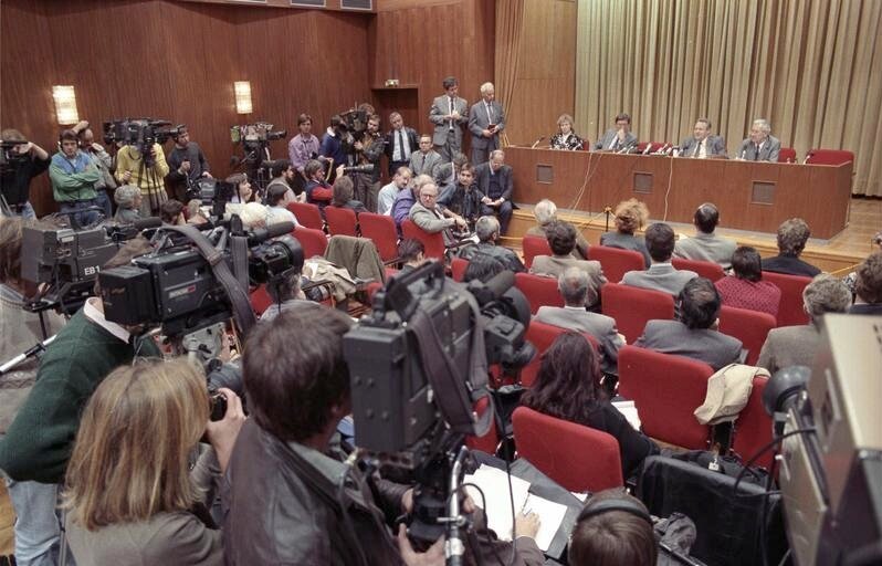 Zdjęcie przedstawia salę, na której odbywa się konferencja. Za długim stołem zabudowanym z przodu siedzi trzech mężczyzn i jedna kobieta. Wszyscy są w średnim wieku. Mężczyźni ubrani są w garnitury, białe koszule i krawaty, a kobieta ma garsonkę i białą koszulę. Przed nimi na stole ustawione są mikrofony. Z lewej strony, obok stołu, stoi lub siedzi na podwyższeniu, na którym ustawiony jest stół, kliku mężczyzn ubranych w garnitury. Na wprost udzielających odpowiedzi osób, w kilku rzędach, na czerwonych fotelach siedzą pozostali uczestnicy konferencji. Są wśród nich kobiety i mężczyźni ubrani w oficjalne, eleganckie stroje. Trzech mężczyzn z pierwszego rzędu odwraca się do tyłu, przysłuchując się pytaniom zadawanym z głębi sali. Za siedzącymi oraz z boku sali stoi wielu przedstawicieli telewizji i prasy. Wyposażeni są w kamery i mikrofony. Dziennikarze mają mniej sformalizowane stroje: swetry, koszule, bluzki, kurtki.