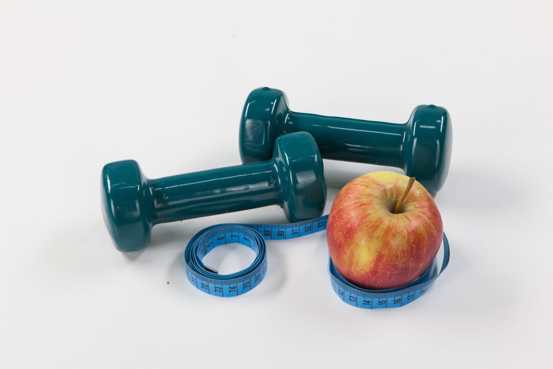 Fotografia przedstawia dwa niebieskie ciężarki do ćwiczeń, błękitny centymetr krawiecki i czerwono – żółte jabłko z ogonkiem. 