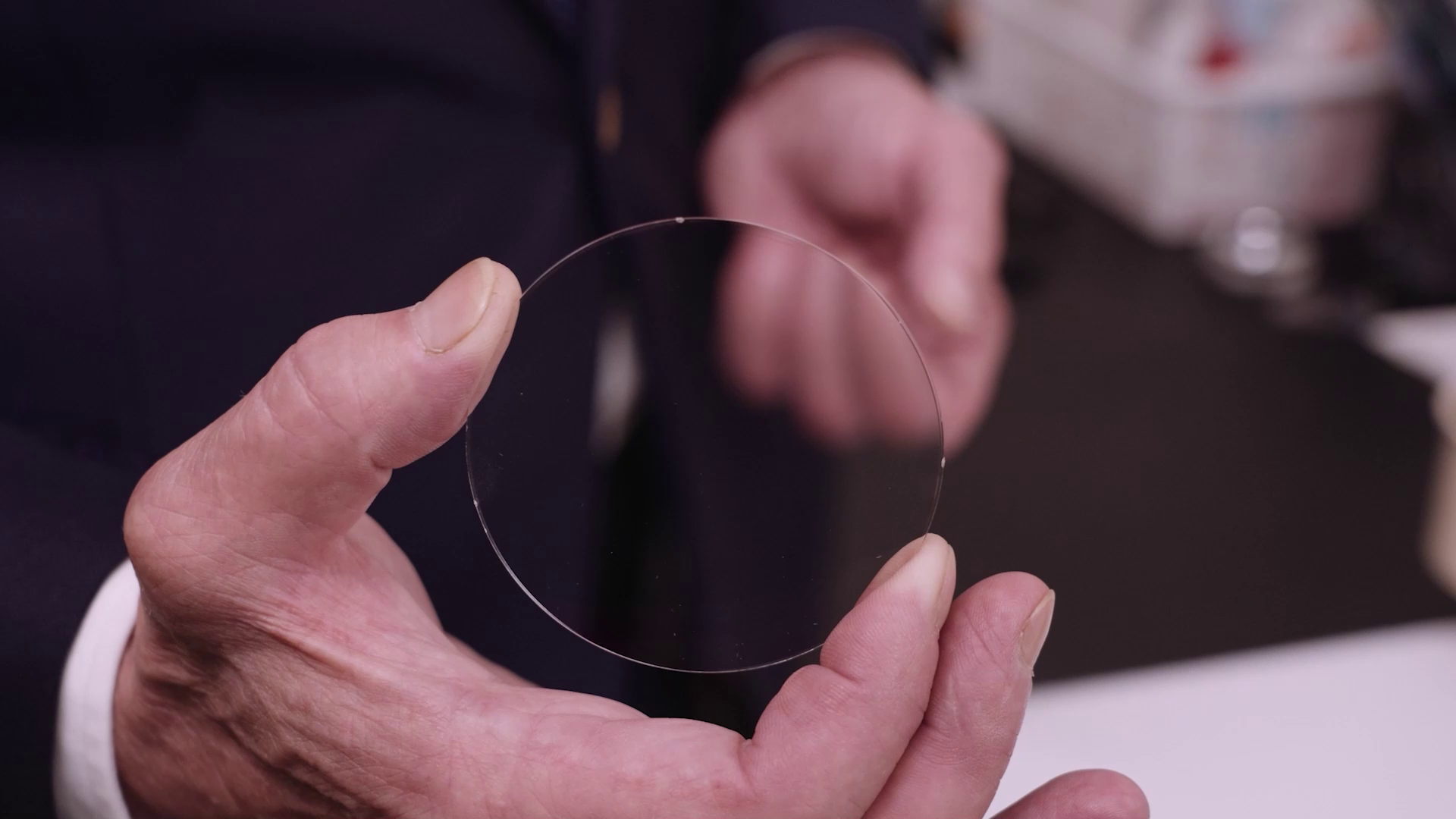 Grafika przedstawia zbliżenie na okrągłe i przezroczyste szkło optyczne trzymane w dłoni mężczyzny.