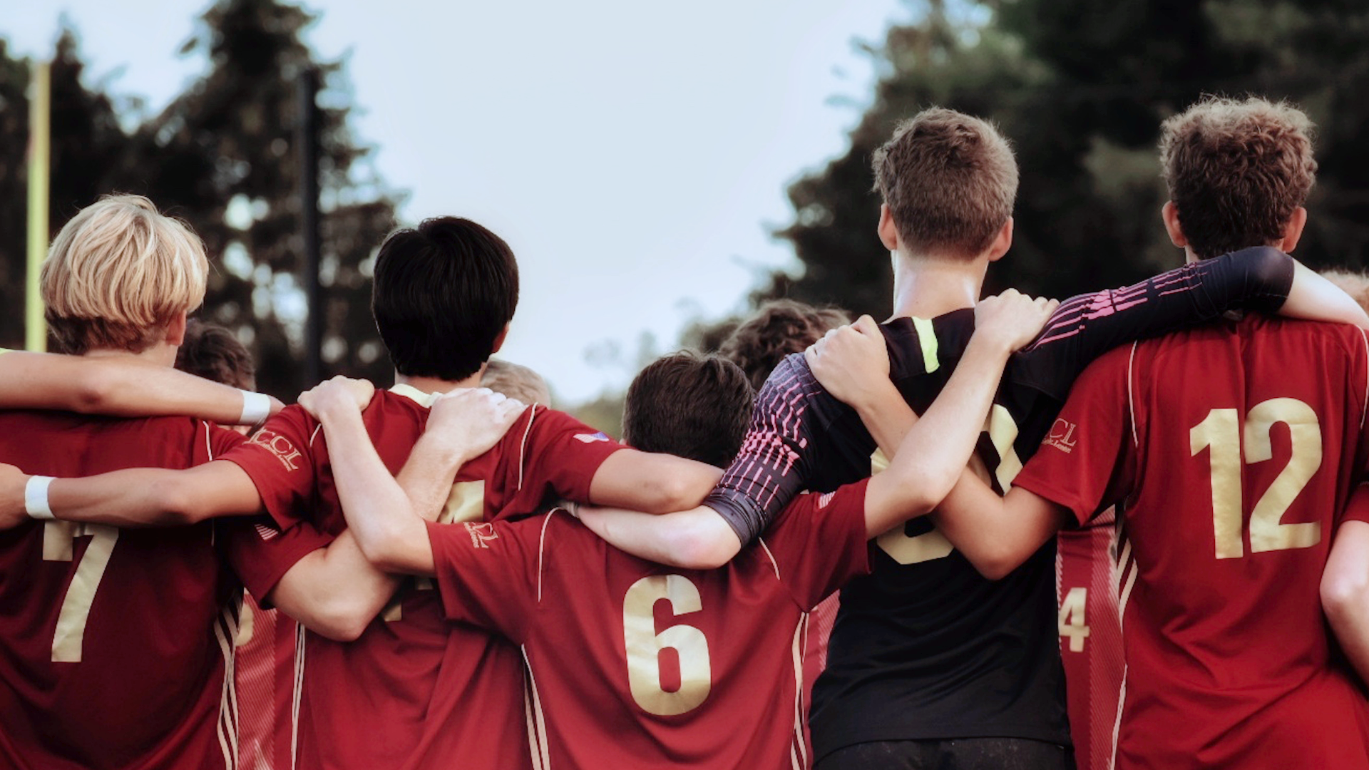 Zdjęcie przedstawia pięciu młodych piłkarzy stojących tyłem do kadru. Piłkarze stoją w rzędzie i obejmują się trzymając ręce na ramionach.