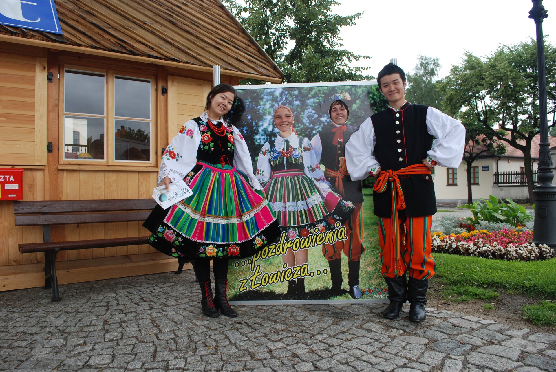 Zdjęcie przedstawia dwójkę Azjatów w polskich strojach ludowych na tle drewnianej chaty.  