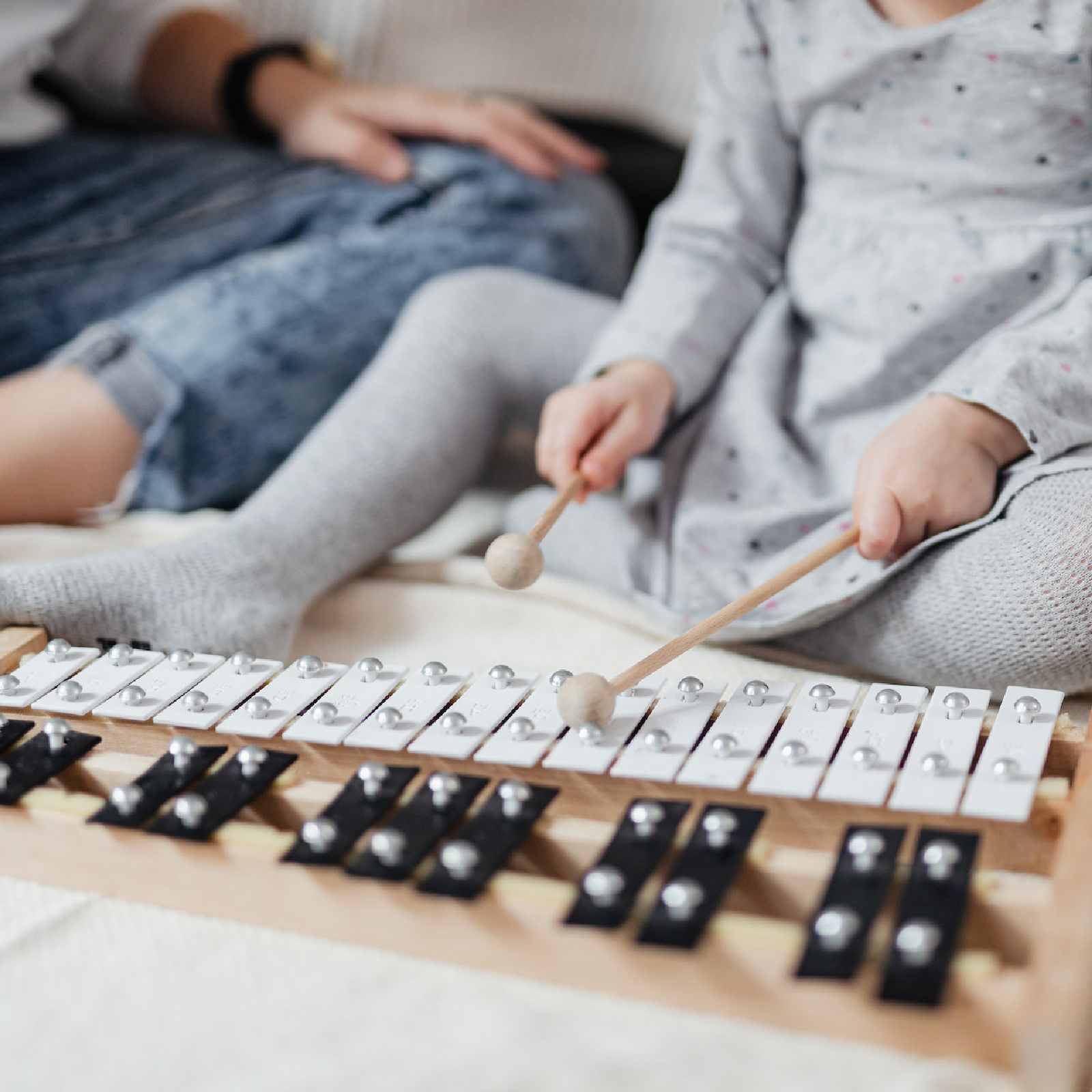 Ilustracja przedstawia biało‑czarny ksylofon. W tle widać osobę dorosłą oraz dziecko, które uderza pałeczkami w instrument. 