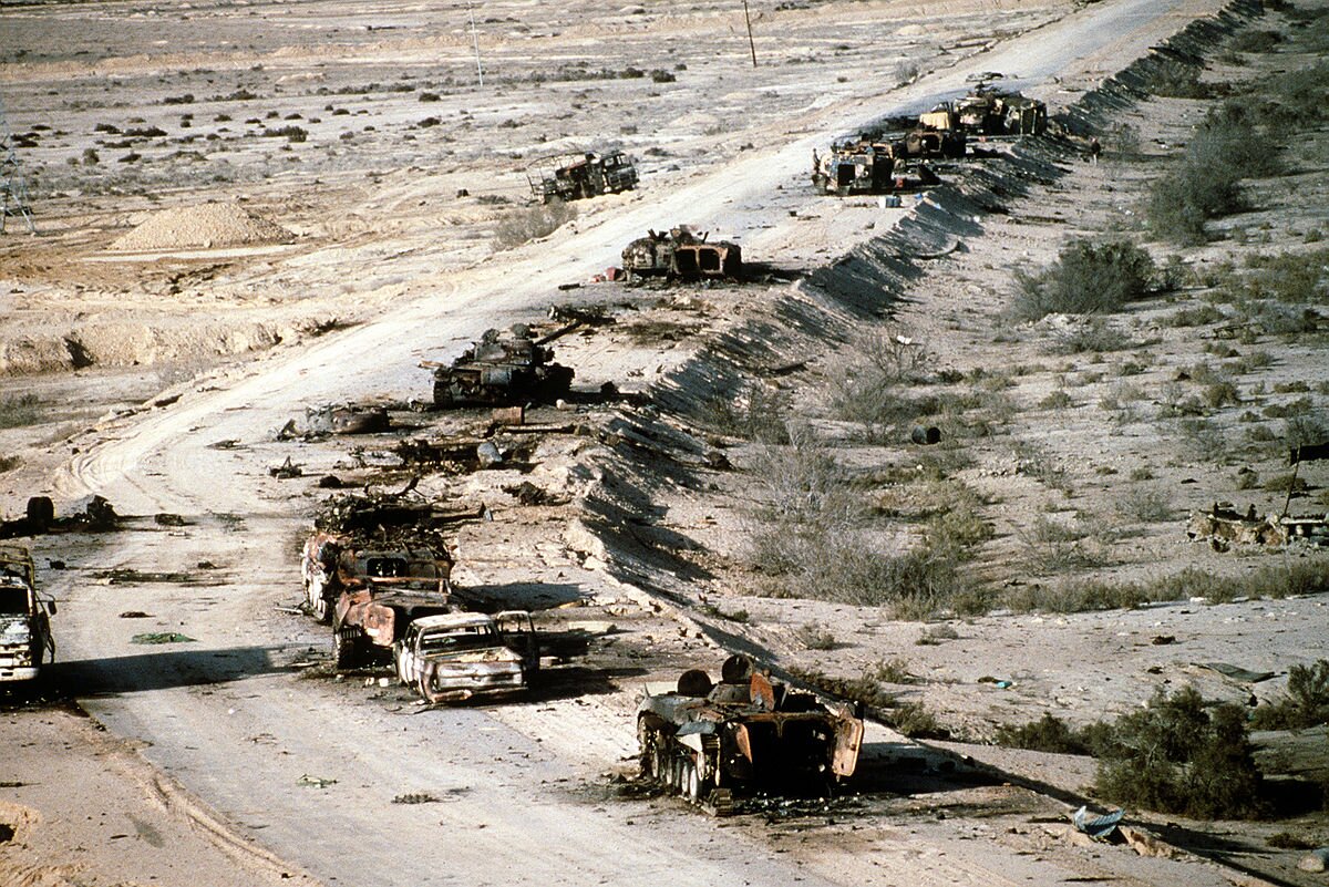 Zdjęcie przedstawia drogę prowadzącą przez pustynię. Na drodze stoją zniszczone, spalone transportery opancerzone, czołgi i ciężarówki.