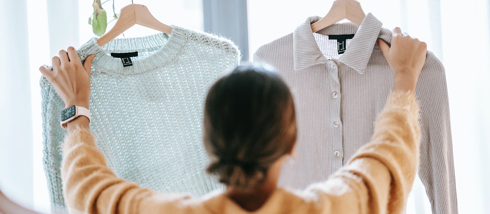 Zdjęcie przedstawia kobietę, w ujęciu od tyłu od ramion w górę, która w rękach uniesionych do góry trzyma dwa drewniane wieszaki. Na lewym jest powieszony niebieski sweter, a na prawym jasna koszula. 