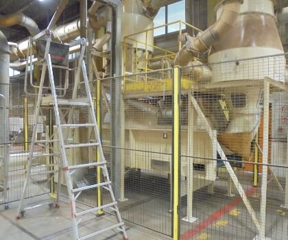 Zdjęcie przedstawia opener - maszynę do produkcji wyrobów z włókna kokosowego. Stoi w hali zakładu produkcyjnego. Jest zabezpieczona ogrodzeniem z siatki. Urządzenie połączone jest z rurami o różnej średnicy i posiada dwie komory w górnej części. 