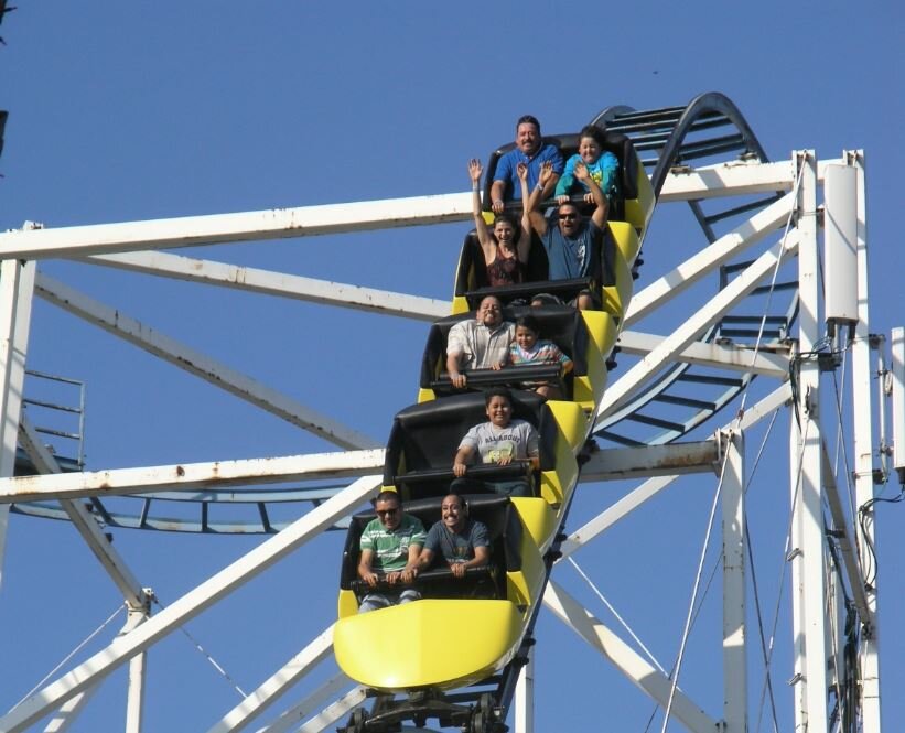 Zdjęcie przedstawia na tle niebieskiego nieba biało szarą ażurową konstrukcję z wagonikiem, w którym są ludzie. Dwie osoby mają uniesione ręce.