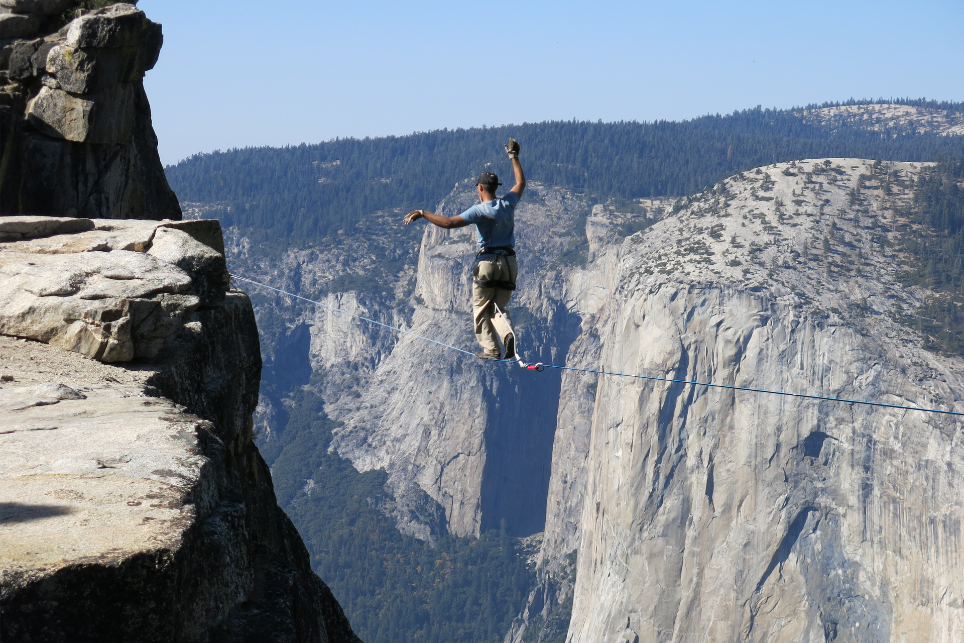Fotografia przedstawia człowieka idącego po linie nad przepaścią w górach. Przy pomocy uniesionych rąk człowiek utrzymuje równowagę. Ma też linę zabezpieczającą.