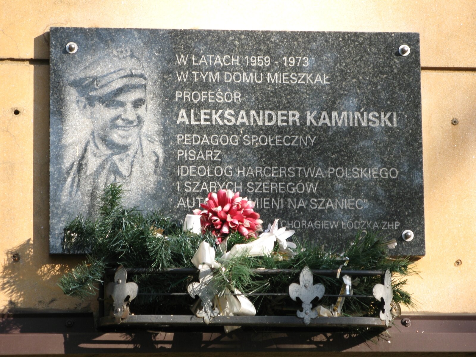 Zdjęcie tablicy pamiątkowej z czarnego kamienia, która jest umieszczona na ścianie budynku. Po lewej stronie znajduje się podobizna pisarza, po prawej napis: „W latach 1959‑1973 w tym domu mieszkał profesor Aleksander Kamiński, pedagog społeczny, pisarz, ideolog Harcerstwa Polskiego i Szarych Szeregów, autor „Kamieni na szaniec" Chorągiew Łódzka ZHP". Pod tablicą ułożone są sztuczne gałązki świerkowe i czerwony kwiat.