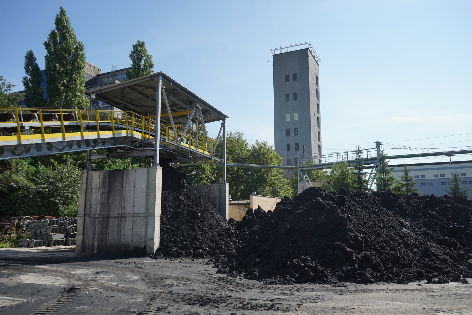 Na zdjęciu widoczne jest składowisko z węglem. Widoczne na zdjęciu zwały mają kształt pryzm. Nad pryzmami widoczny jest podwieszony przenośnik taśmowy. Budynek przypomina cienki prostopadłościan. W tle znajdują się budynki przemysłowe.Poniżej zdjęcia widoczny jest przycisk multimedialny z nagraniem audio. Treść nagrania jest zgodna z tytułem zdjęcia.