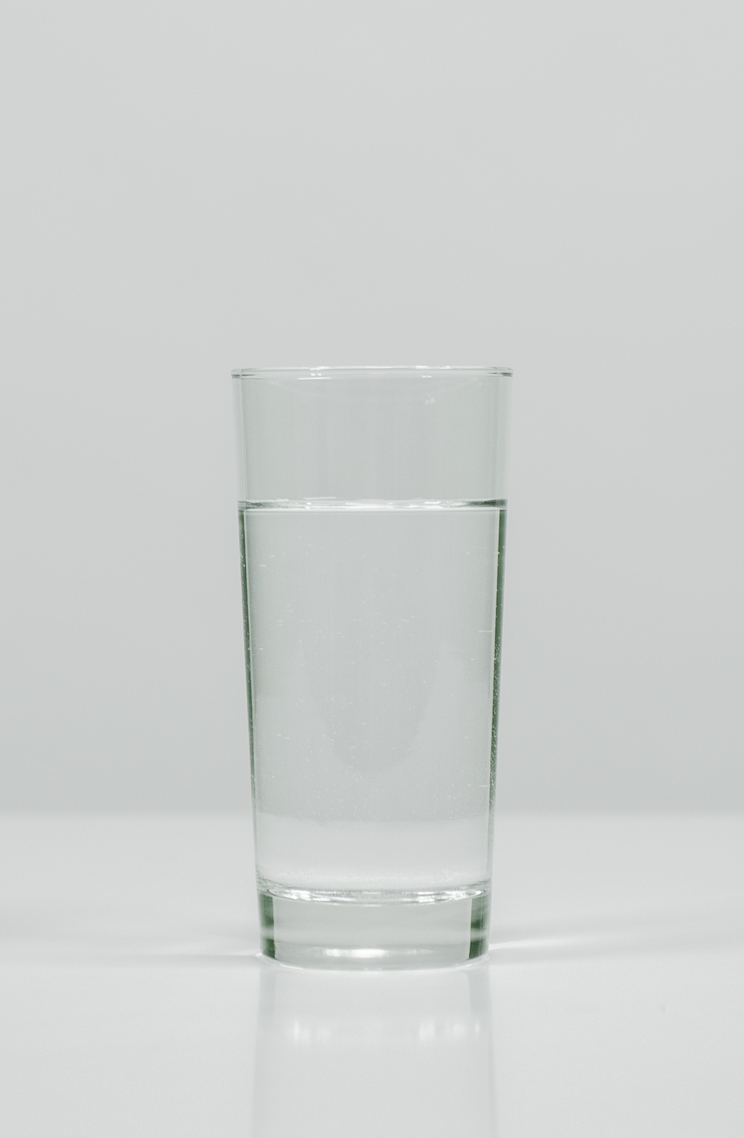 Zdjęcie przedstawia szklankę z wodą stojącą na płaskiej, jasnej powierzchni. Zawartość szklanki jest klarowna, woda wygląda na idealnie czystą.